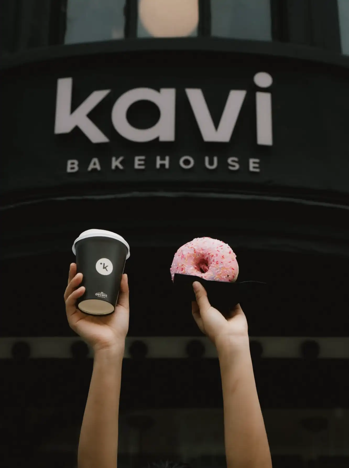 a takeaway coffee and a pink glazed doughnut held up outside kavi bakehouse